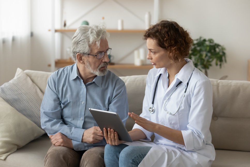 Een dokter zit op de bank met een oudere man terwijl ze bespreken wat er op een tablet staat