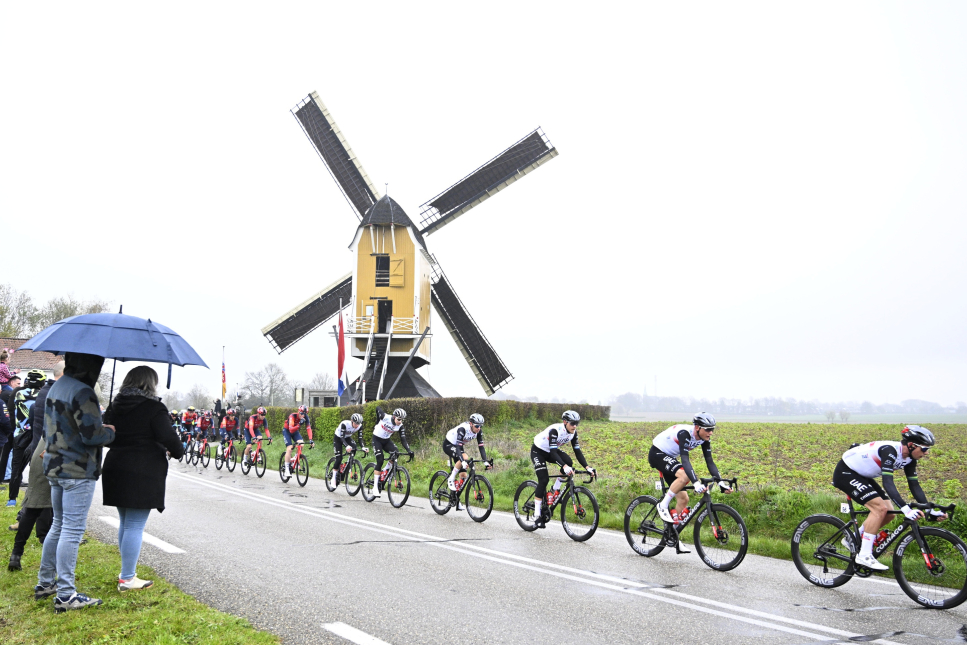 Mensen onder een paraplu kijken hoe wielrenners tijdens de Amstel Gold Race van 2023 door een regenachtig Limburgs landschap koersen.