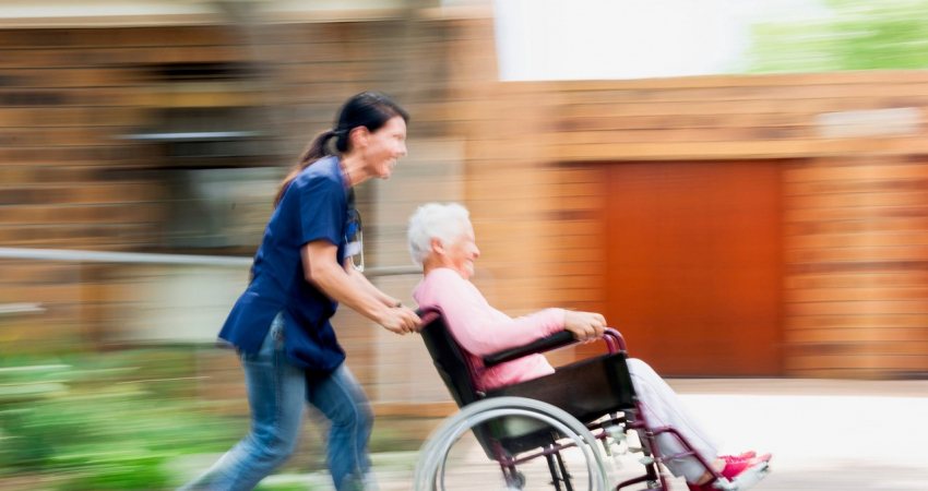 Een verzorgster rent met een oudere vrouw in rolstoel over de straat