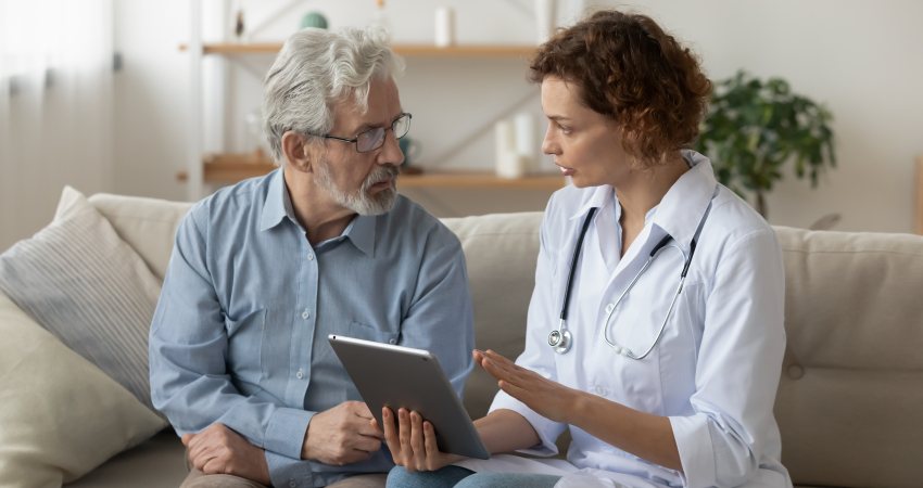 Een dokter zit op de bank met een oudere man terwijl ze bespreken wat er op een tablet staat