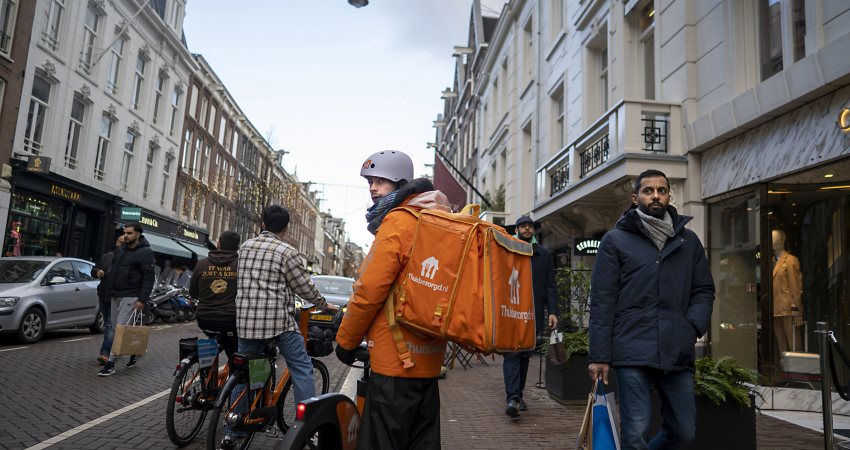 Een maaltijdbezorger staat met fiets en rugzak tussen het winkelend publiek in Amsterdam. 