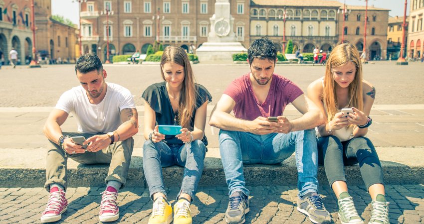Vier jongeren zitten aan de rand van een plein op hun mobiele telefoon.