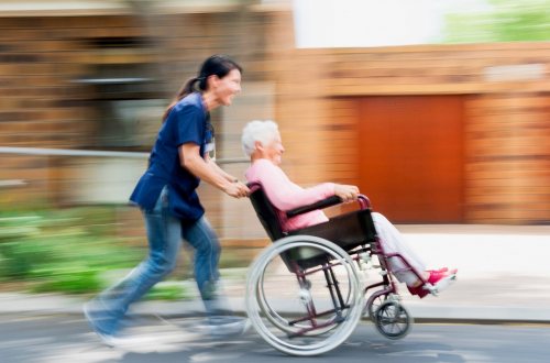 Een verzorgster rent met een oudere vrouw in rolstoel over de straat