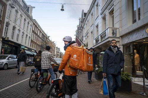 Een maaltijdbezorger kijkt vragend achterom in een Amsterdamse straat terwijl het winkelend publiek aan hem voorbij trekt.