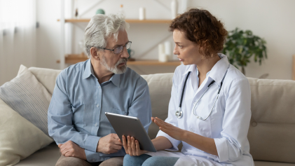 Een dokter zit op de bank met een oudere man terwijl ze bespreken wat er op een tablet staat