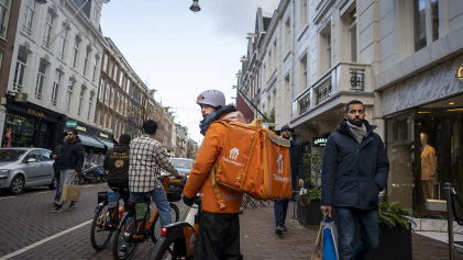 Een maaltijdbezorger kijkt vragend achterom in een Amsterdamse straat terwijl het winkelend publiek aan hem voorbij trekt.
