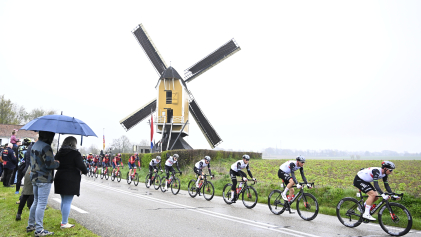 Mensen onder een paraplu kijken hoe wielrenners tijdens de Amstel Gold Race van 2023 door een regenachtig Limburgs landschap koersen. 