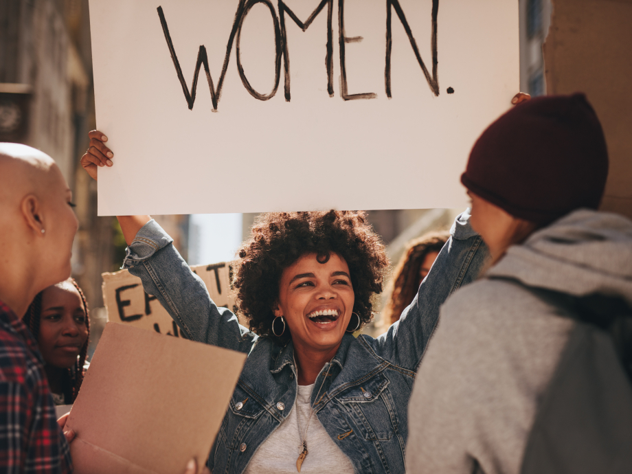 Een vrouw houdt lachend een bord omhoog met 'women."