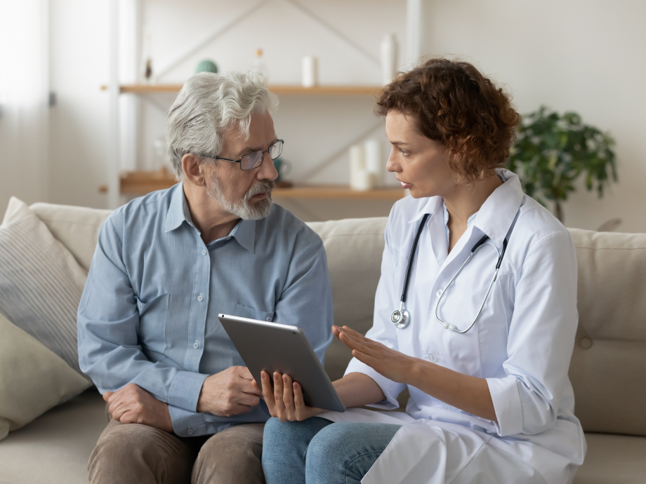 Een dokter zit op de bank met een oudere man terwijl ze bespreken wat er op een tablet staat