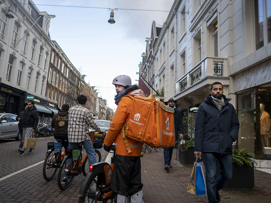 Een maaltijdbezorger kijkt vragend achterom in een Amsterdamse straat terwijl het winkelend publiek aan hem voorbij trekt.