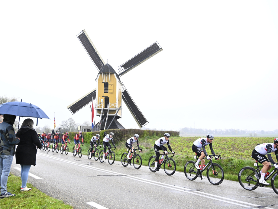 Mensen onder een paraplu kijken hoe wielrenners tijdens de Amstel Gold Race van 2023 door een regenachtig Limburgs landschap koersen. 